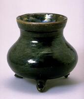This black glazed vessel stands raised on four feet and has a high neck.