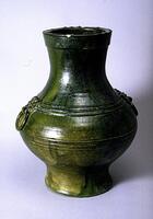 This tall, red earthenware jar with bulbous body, curved flaring neck, and direct rim with articulation rests on a tall, slightly flared foot ring. Appliqué "bow string" bands wrap around the body and rim, and two animal mask decorations hold rings on opposing sides of the belly. The jar is covered in a dark green lead glaze.