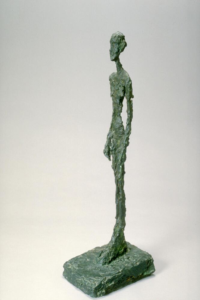 An elongated female nude figure with minimal features stands on a small rectangular base with left foot slightly forward. The metal has a texture resembling molten wax.