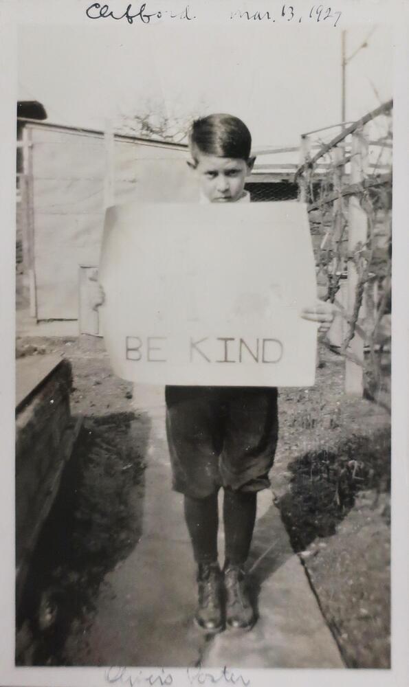 A boy holding a sign that reads &quot;BE KIND&quot; in capital letters. His face is turned down slightly with his eyes looking up at the camera.