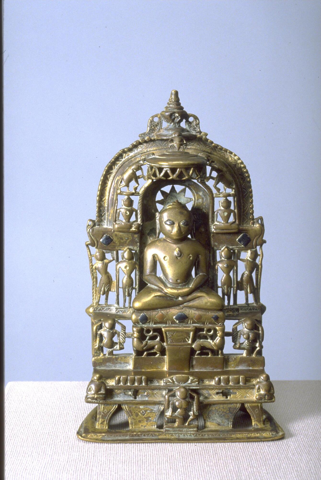The jina Malli sits in the lotus position on an inlayed cushion on a tiered throne. Seated with his hands folded in a gesture of meditation, he is surrounded by a number of figures representing other jinas, attendants and demigods. In the center in front of the throne sits the goddess Ambika with a child on her lap.  On the first tier of the throne sit two figures that may represent donors.  On the next left are nine mounds representing the nine planets [navagraha], five to his right and four to his left.  At the base of his seat are two stylized lions and this is flanked by a male and female demigod.  On the arch surrounding the figure at his level a standing jina figure is to each side and cauri bearer is on the outside of each of them.  At his shoulders, the cross bars of the throne back end in stylized makara heads with jewels hanging from their mouths.  A seated jina adorns the arch to each side of his head and elephants surmount them with an umbrella over his head with a dancing figure atop it.  The who