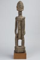 Male figure with bent knees holding an L-shaped tool. The figure is mounted on a wooden base.