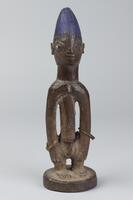 Standing human figure on a round base with hands at the sides. The torso and arms are cylindrical and around the waist is a string of beads. The hair is conical in shape with a rounded top and has incised, vertical grooves. 