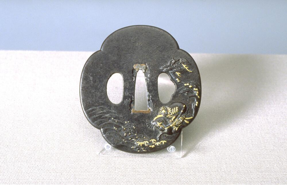 This small, flat metal piece has a quartrefoil shape. Three holes in the middle. Some chips can be seen around the center hole, which mended with silver and copper. A samurai on horseback is charging into the sea from steep hill; he is wearing a helmet, armor, and sword, and holding a fan. A pine tree is standing on the samurai’s right side; there are rocks and bamboo grass by the ocean. The motifs of the ocean, pine tree, bamboo grass and rocks also appear on the reverse side. Gold and some silver and copper inlays are applied on pine branches, samurai's helmet, horse's mane and bridle, bamboo grass on the shore, and spray from waves.