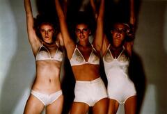 The work is a color photograph of three models in various styles of underwear and brassieres. They stand close to one another, with their arms outstretched above their heads. They stand against a blank wall, their dark shadows projecting behind them.