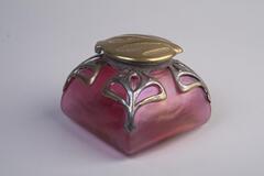 A square, red satin glass inkwell with a flat, brass lid.  Silver designs are applied to the corners.