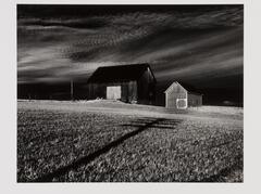 Two barns set perpendicular to one another in a grassy field with a long overcast shadow of a utility pole.