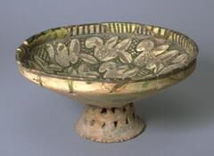 This footed bowl with painted designs is of 11th-13th century Iranian origin. The object stands upon a broad pierced foot, with a stem extending upwards supporting a shallow dish. It is done in brown, green and gold. The decoration consists of a number of birds compartmentalized by a gold background that serves to set off their shapes and creates a free-form design. Sgraffito cuts through the slip on glaze to a layer of brown that creates part of the form of the bird or its decoration. All of this is set against green cross-hatched areas. <br /><br />
 
