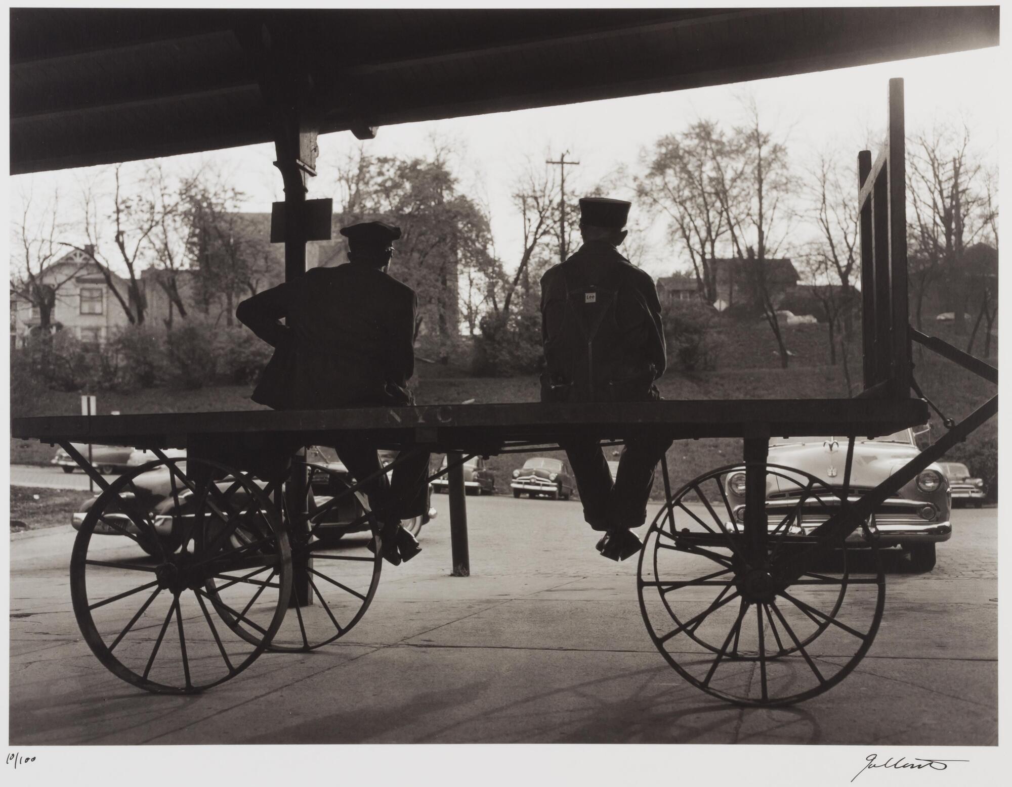 Two men seated on part of a railroad car at a station. A parking lot is in front of them.