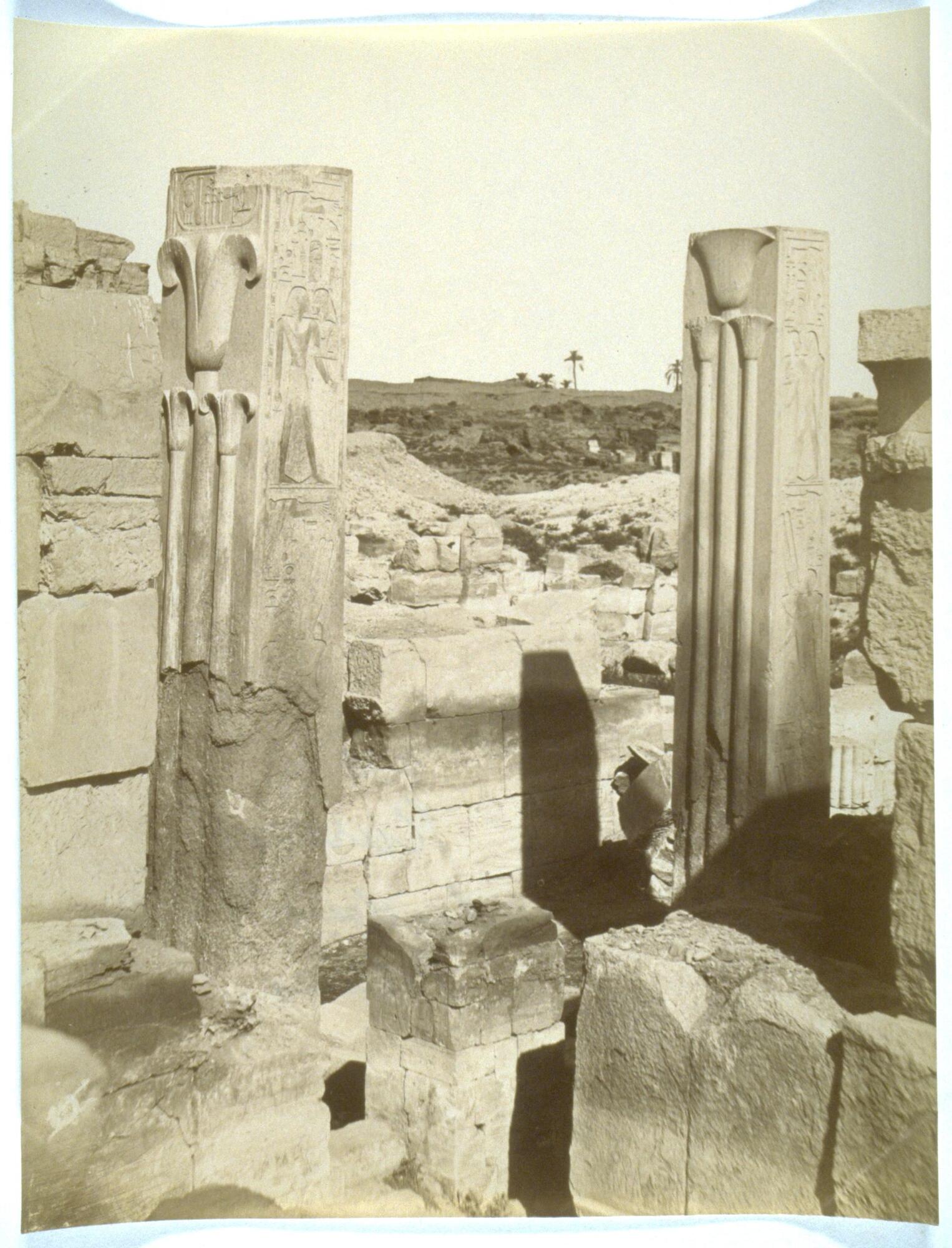 This photograph depicts a view of some ancient stone ruins. Two columns stand in the center of the photograph, each bearing relief designs and carved hieroglyphics.