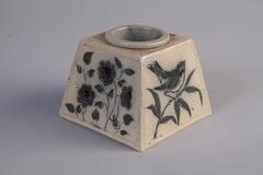 A square ceramic inkwell with a motif of a bird on a branch on one side and a motif of flowers on the other. The base of the inkwell is beige and the pictures are green and black.