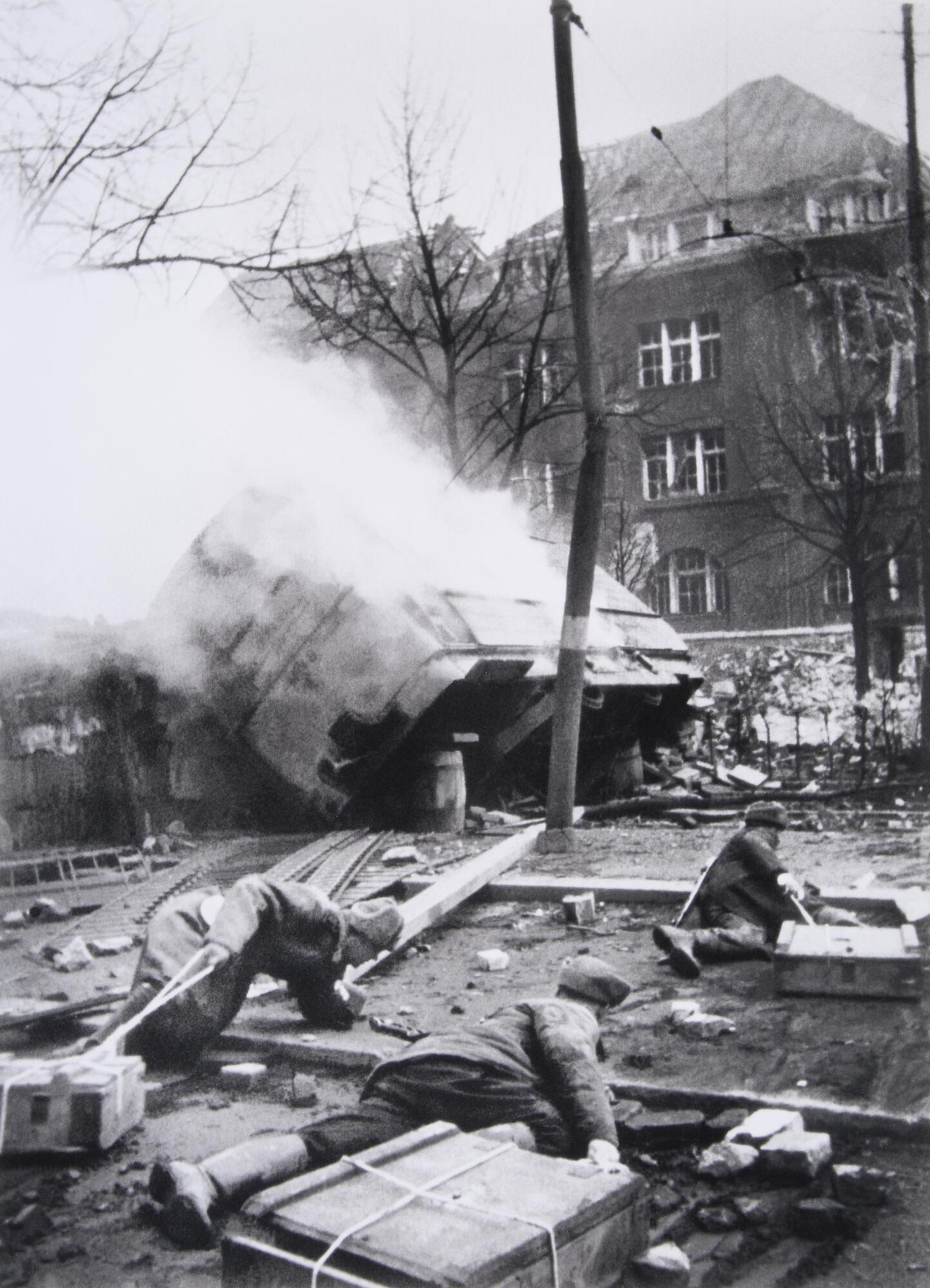 Three soldiers crawl across a city street littered with rubbled and burning vehicles while towing crates of supplies. 