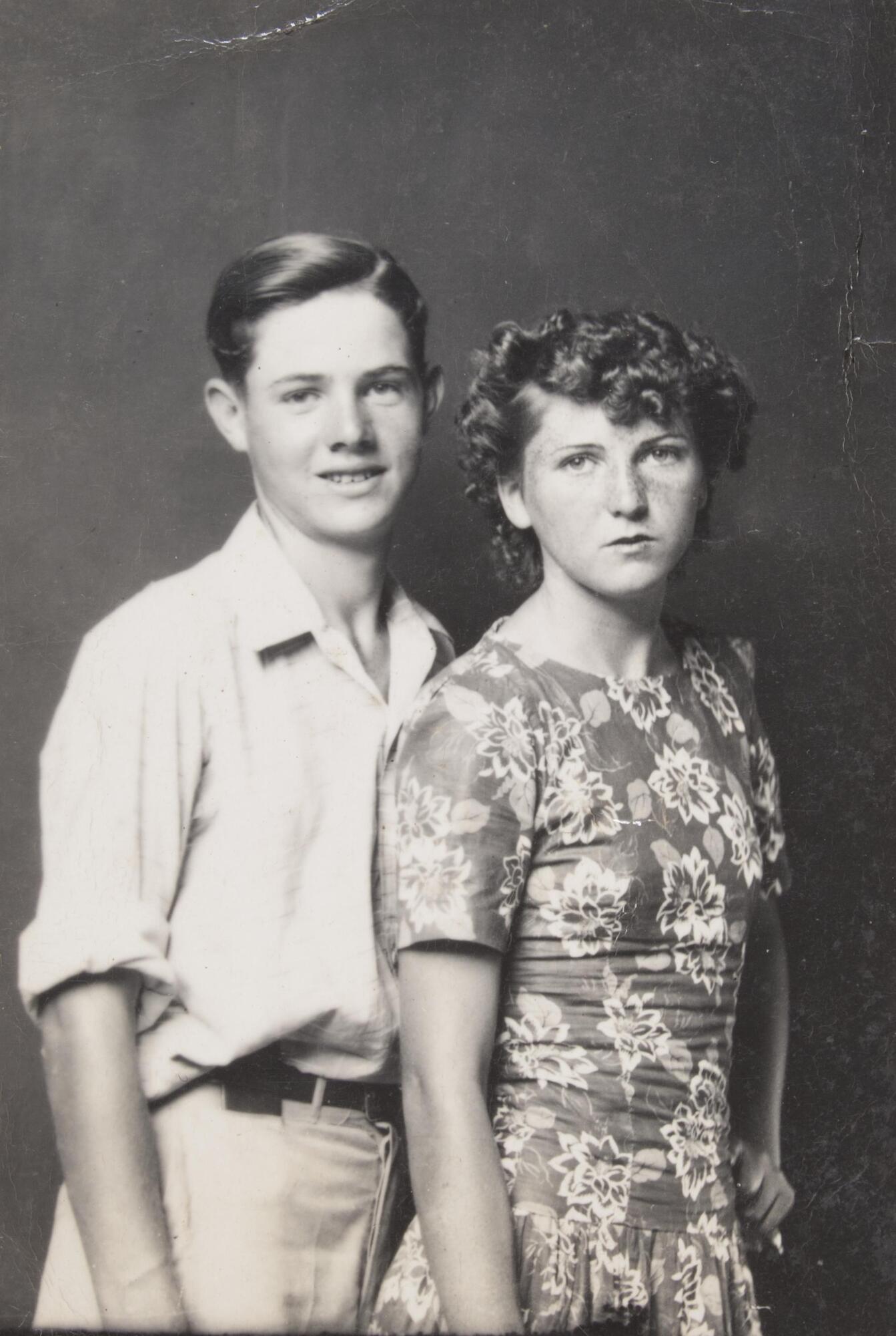 A portrait of a young man and woman standing next to one another. The young man wears a shirt, belt, and trousers. The young woman wears a floral dress.