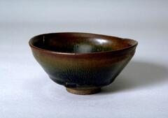 A deep, conical bowl on a straight foot ring and subtle rim articulation, covered in a thickly applied dark iron-rich black glaze with lighter russet-brown hare's fur (兔毫盏 <em>tuhao zhan</em>) markings.  The thick glaze thins at the rim to a russet-brown color and pools to black near the foot ring.
