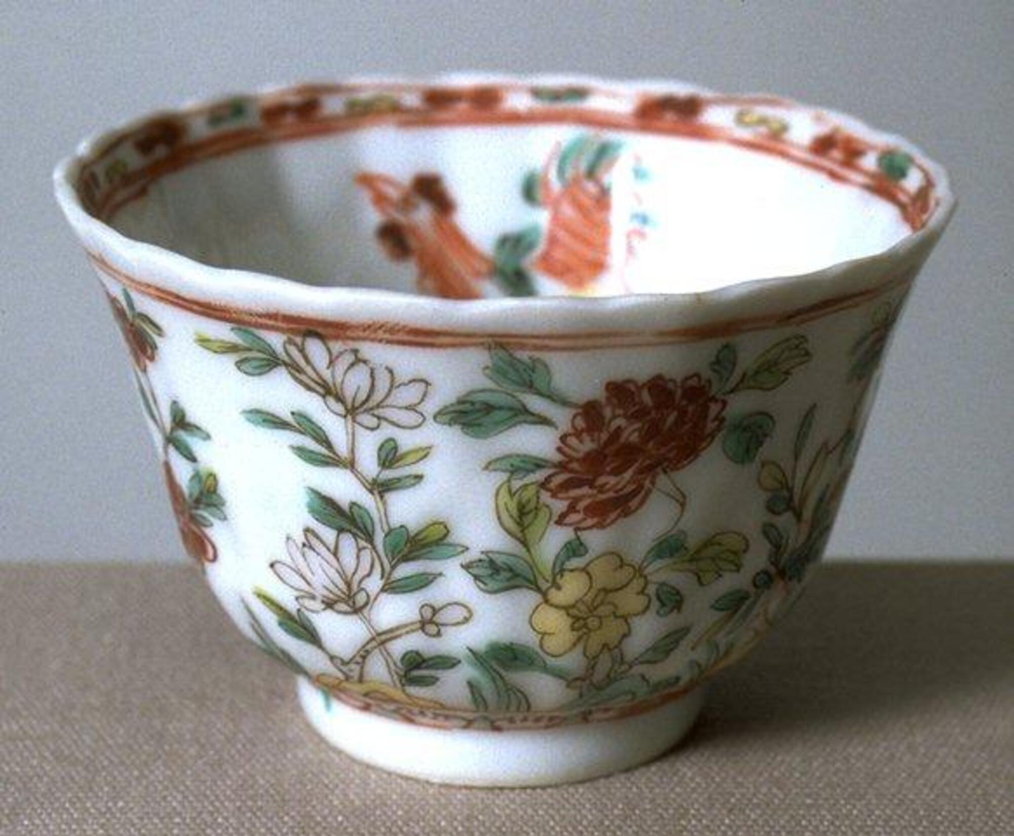 This bowl flares out by having a smaller diameter on the bottom and larger diameter along the rim. The outside of the bowl is decorated with red, pink, and yellow peonies with green leaves. The inside of the bowl is decorated with a red rooster. The outside rim of the bowl has two red horizontal stripes, the top rim of the bowl is wavy with many bumps that resemble flower petals, the inside rim is decorated with more peonies. The foot of the bowl it tall.