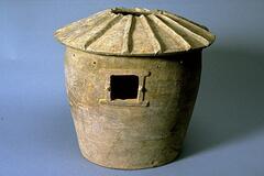 A gray earthenware cylindrical granary, with a framed, pierced window, and a round hipped and peaked roof with a central opening and spoke-like roof ridges. The model is covered in a lead green glaze, with iridescence and calcification. 