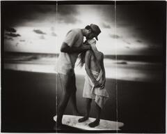 Man leans over to kiss the forehead of a young girl standing on a surfboard on the beach.