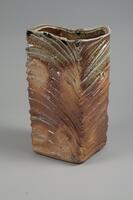 The exterior of this three-sided triangular based vase is decorated with lines that curve together from one edge of the vase to the next. The ash glaze creates a greenish brown color, which pools and creates darker tones in some areas. The edges are not straight, but rough and bumpy.