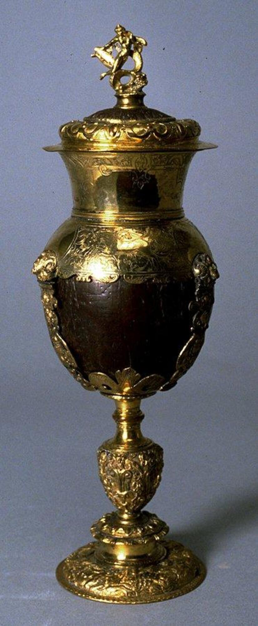 This cup is fashioned from a coconut shell set in a shallow bowl supported on a long, narrow stem and held in place by three straps and a tall neck band. The shallow domed lid has a plain, overhanging rim and terminates in a finial with a statuette of a nude sea nymph and serpent. The metalwork is densely decorated with various masks, animals, fantastic creatures, and vegetal ornament.
