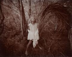 In this photograph of a young girl she stands in a wooded area. She stands in between small trees, looking up, with the long skirt of her dress wrapped around her arms.
