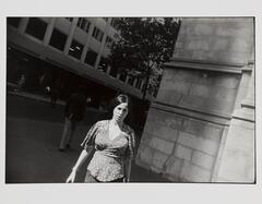 Photograph of a woman walking on a sidewalk next to a building; she emerges from the shadow into the sunlight.