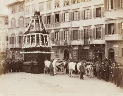 This photograph depicts a procession of a large, ox-drawn cart covered in flowers in an Italian city. Gathered around the cart are a group of dark-suited men.  
