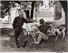 Two men and a dog. The dog has a paw on the one man's leg while he is petting it. The other man is standing with his elbow leaning on a mailbox.