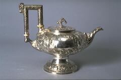 Short, squat silver pot with short spout, finial-topped lid, tall square handle and opulent repouss&eacute; decoration