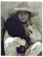 This is a photograph of a young boy in Hidalgo, Mexico. The boy wears a straw sun hat, and crouches on the ground with his left hand on the side of his face. Draped over his right shoulder is a cloth blanket or poncho.