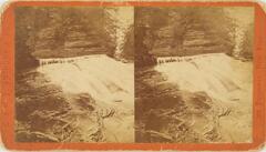 This black and white stereoscopic image features two images of a small water fall that flows from the top left to the bottom right. There is a tall wall of rock towards the top and flat smooth rocks with water flowing over them in the bottom left corner. This image is a sepia tone mounted on a red orange background.
