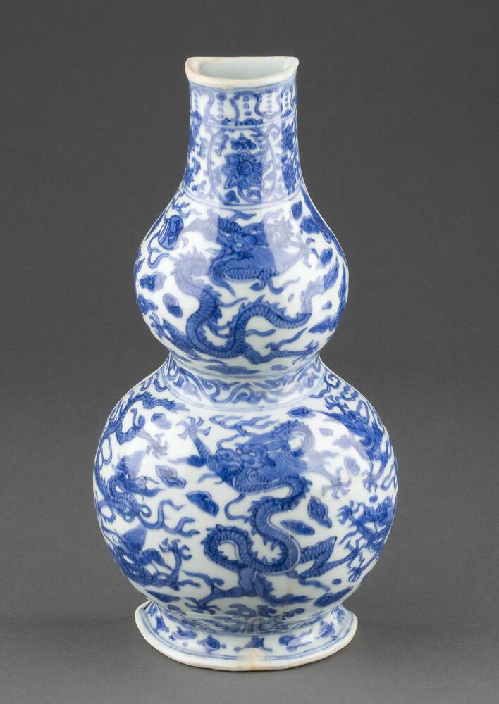 A porcelain bottle vase of double gourd form on a flared footring with a tall, narrow neck; the reverse side is flat with a slot for hanging. The vase is outlined in underglaze blue and has a six-character Wanli mark in a plaque. The front is decorated with underglaze blue five-clawed dragons among clouds—five dragons on the lower bulb and one on the upper—confined between floral borders. The vase is covered in a clear glaze. 