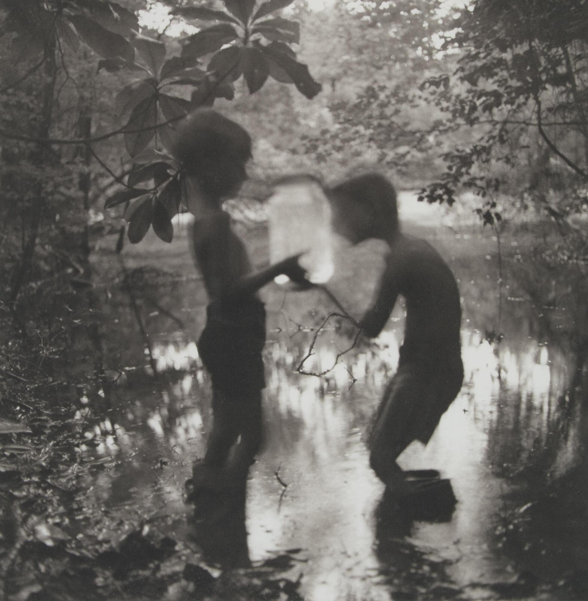 This photograph depicts a view of the silhouetted forms of two boys holding a large glass jar and standing in a body of fresh water surrounded by trees.
