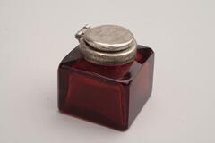 A square, ruby colored inkwell. The lid is made out of a flat piece of metal.