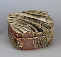 This ovular box, although flat on the bottom, has a lid decorated with curved ridges and protrusions that arch across the surface. The entire exterior is speckled with tiny craters, breaking up the illusion of a perfectly smooth surface. The ash glaze decorates the lidded box with an earthy brown color. The glaz pools in the valleys of the lid and small craters throughout, creating areas of darker hue. The meeting of the lid and box body is not a straight line, but rugged.