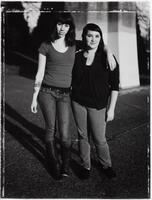 This photograph depicts two women standing side by side on an asphalt-paved street. The taller woman has a tattoo on one arm and affectionately wraps her other arm around her companion’s shoulder.