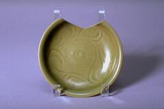 A stoneware wide shallow dish with an everted rim on a footring.  The interior is combed and incised with floral meander, and it is covered in an olive green celadon glaze.  There is loss to the rim and side.  