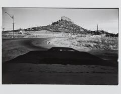 This photograph depicts a view of a landscape with streets lined with telephone poles and a rocky butte rising in the distance. The large, dark shadow of a car is cast on the road in the foreground.