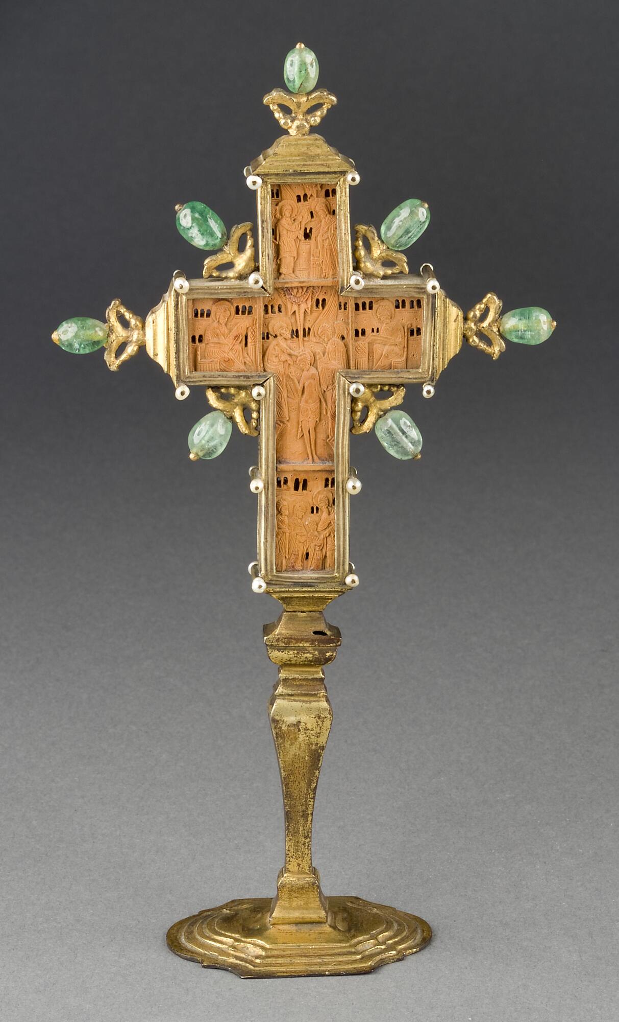 This small carved boxwood cross is divided on each face into five compartments containing Christian religious scenes. The cross is set in a stand decorated with mother-of-pearl and green glass-paste stones.