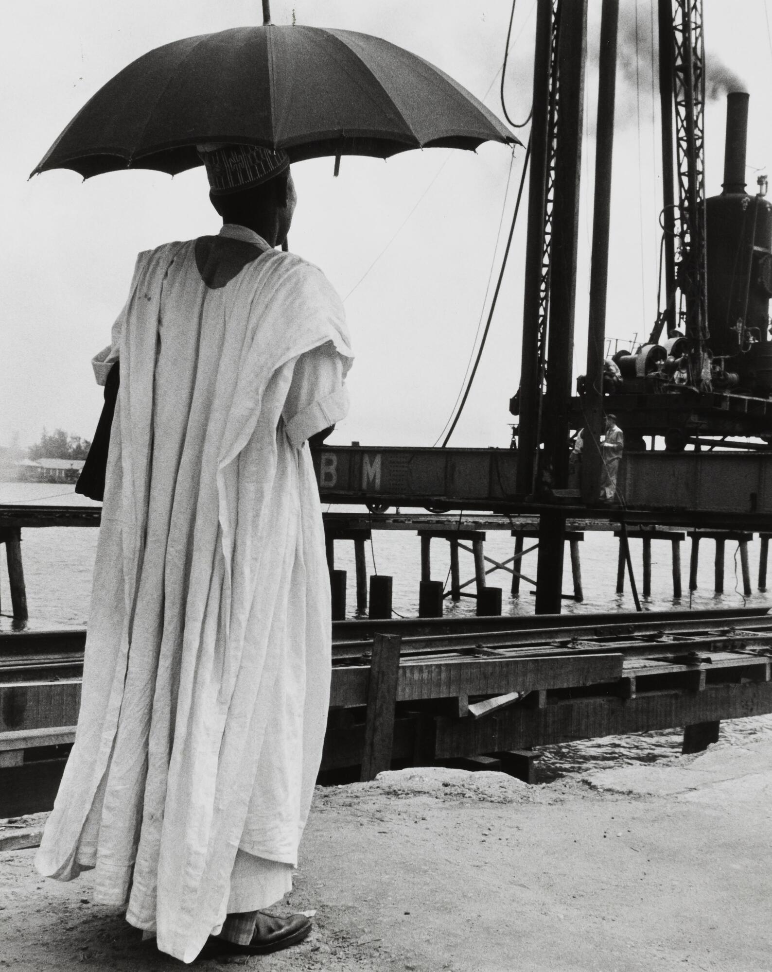A man wearing long, light-colored robes faces away from the camera watching machinery at the edge of a body of water. He holds a dark umbrella.