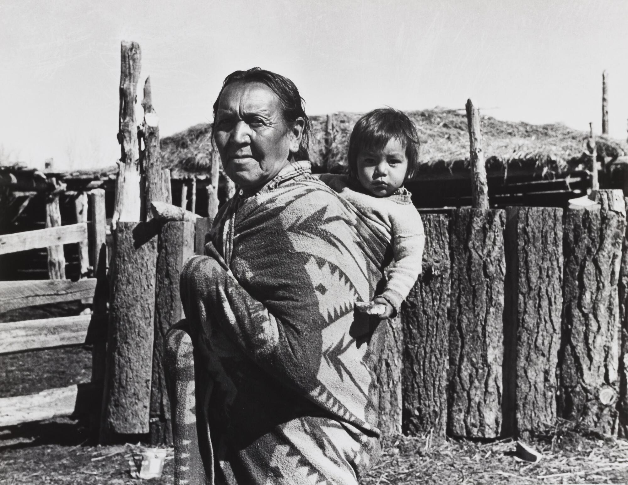 A photograph of an old woman with a baby on her back. She stands in front of a wooden enclosure, fences on the left side of the frame.