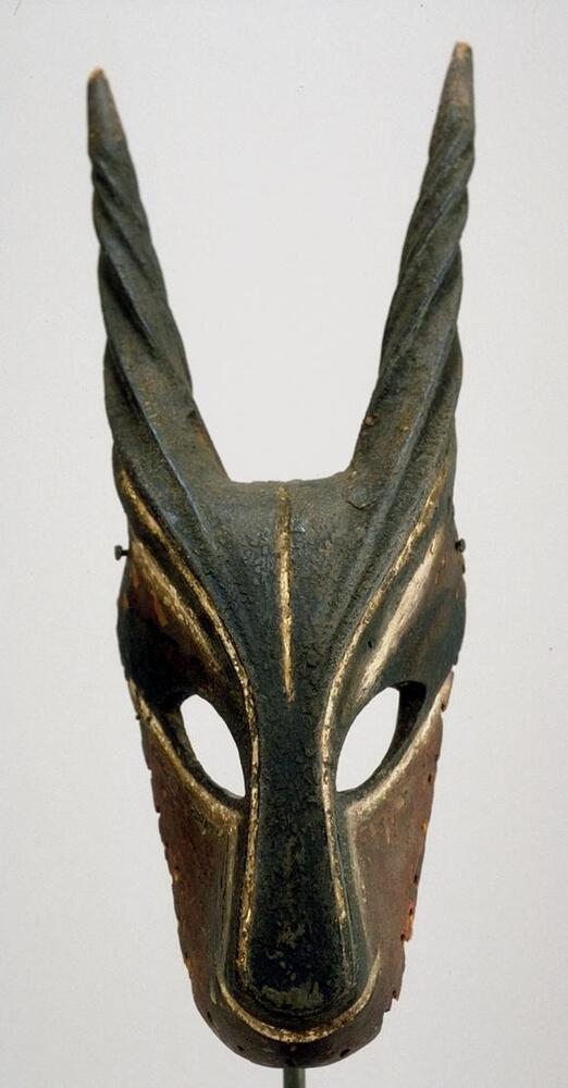 Wooden mask in a triangular shape with rounded edges. There are two almond-shaped holes for eyes and spiral horns protrude from the top of the mask. There are traces of red pigment below the eyes and white pigment outlines the center of the mask.