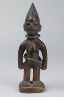 Standing female figure with prominent breasts on a square base. The hands are placed at the sides and there are strings of beads around the waist and neck. On the cheeks and forehead there are incised marks and the hair has a rounded, comb-like shape with horizontal grooves. 