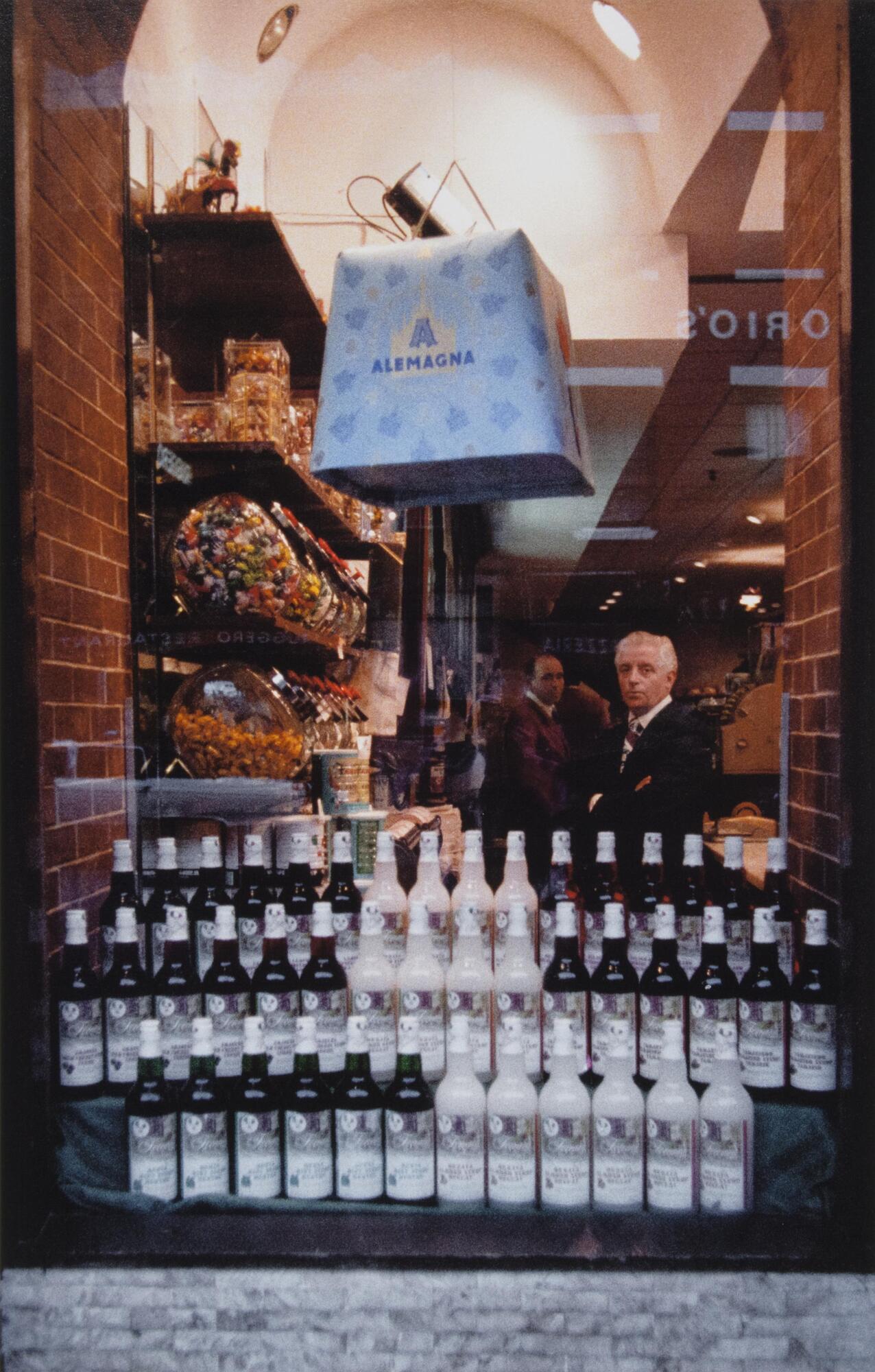 Two men inside a store looking out the window with three rows of bottles in front. "Alemagna" sign is on top of the window.
