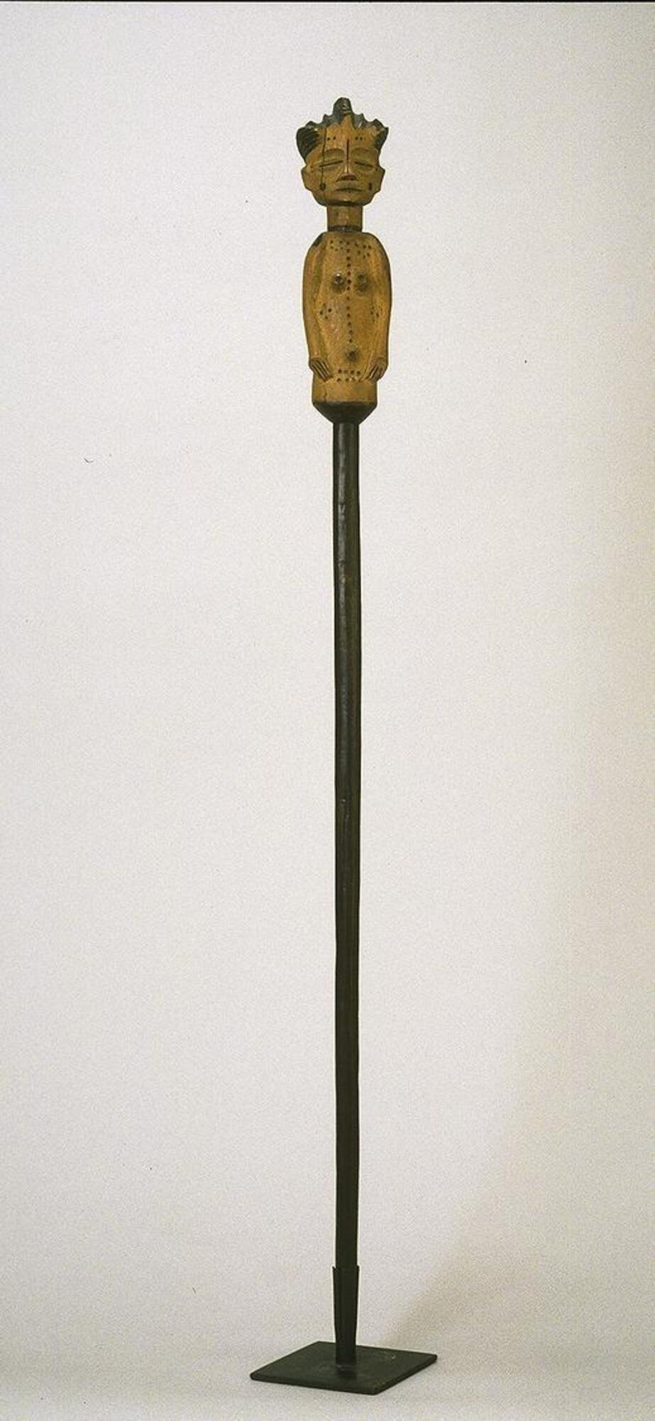 This wooden Chokwe staff features a smooth, narrow rod and a large carving of a female figure at its finial.  The female bears an elaborate, ridged coiffure, closed, coffee-bean shaped eyes, raised scarifications on her face, torso, and back, rounded shoulders, arms positioned down by her side, and a protruding navel. 