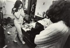 Girl with long hair and hands crossed over body. She is in a messy room and there is a woman with dark hair standing in the foreground.