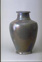 This is a 44.5 cm high ceramic vase. The body has an oval shape with a small rounded band as its base. The shoulder area has a rounded edge and a small flat band at the bottom of the neck. There is a short neck with a rounded, overhanging lip at the top. The vase is covered with an iridescent glaze over a semi-matt glaze that creates an appearance of irregular patches of color. It has a lilac, or lavender blue appearance. The rings of the thrown clay can be seen beneath the glaze.