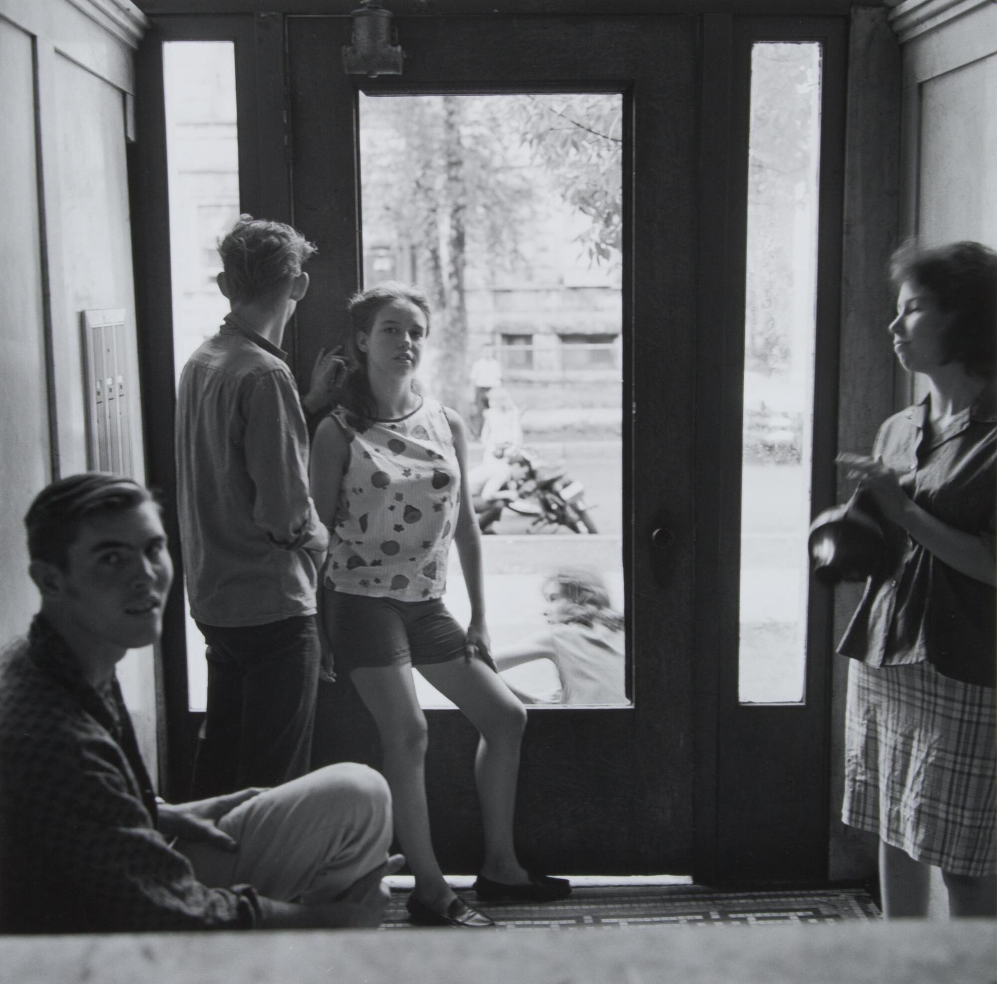 A photograph of two men and two women in an apartment foyer. They stand casually as they interact with one another. Beyond the door, people are visible on the street.