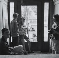 A photograph of two men and two women in an apartment foyer. They stand casually as they interact with one another. Beyond the door, people are visible on the street.