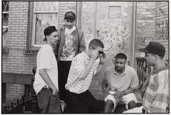 A photograph of a group of five young men in front of a door and brick wall, which are covered in graffiti. Two men stand on the left, one puts on sunglasses and crouches toward the other two, one sitting and one standing, on the right.