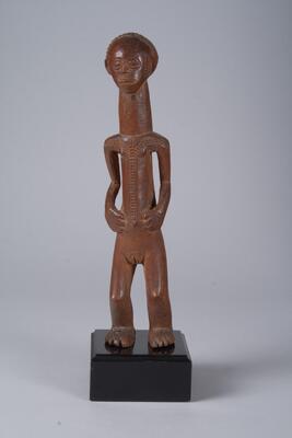 Carved wooden figure of a human. The torso, neck and head make an elongated cylinder along the certical axis. The hands rest at its sides near the umbilicus. The hair, face, hands, genitals, and feet are detailed.